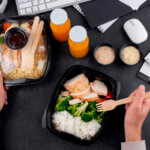 Контейнеры с едой на офисном столе: курица, рис, овощи, соусы и соки